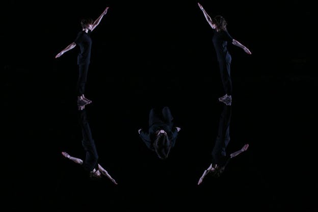 Two performers dressed in black stand symmetrically facing each other and raising one arm in front and one in back with one performer lying with their back on the floor between them in a black room with a mirrored floor reflecting their bodies. 