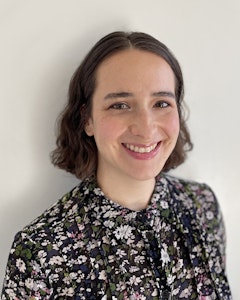 Portrait of a smiling Anastacia Bloch with short brown wavy chin-length hair parted in the middle, dressed in a floral dress shirt with colors of pale pink, blue, and green.