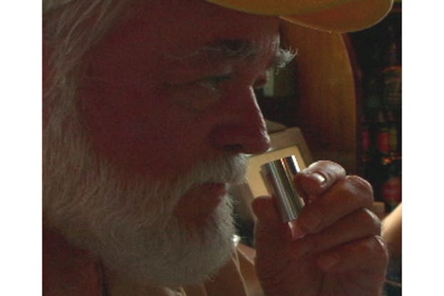 Close-up of a white-bearded man smelling a silver cylinder. 