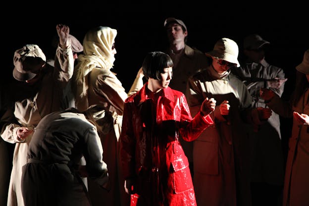 Nine performers stand in various postures as if they were holding the straps and poles of a subway. They wear long coats, hats, and scarves. A single figure is illuminated in the center of the image wearing a bright red coat and looking to the right.