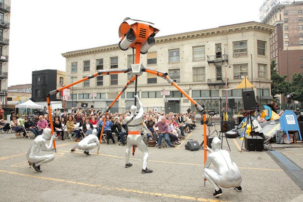 Four performers in silver leotards hold the legs of an orange spider-like machine as a seated audience watches. 