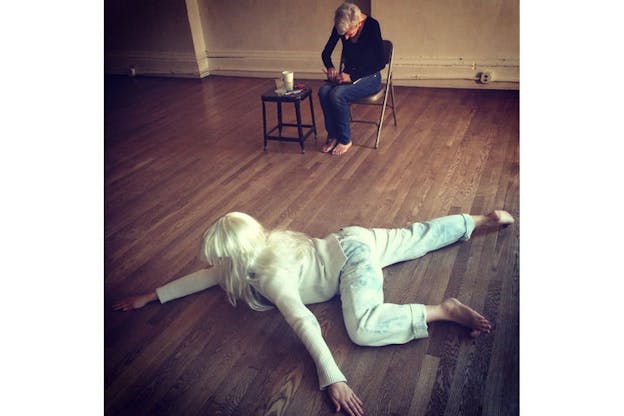 Performer, barefoot in a white sweater and light jeans, sprawled on their stomach on a wooden floor. Behind, a person siting on a chair, working on something in their lap.