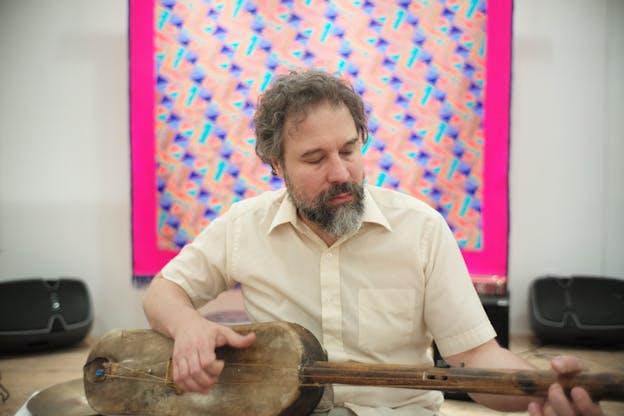 A person dressed in a yellow button up en face to the viewer, looks down on a string instrument as he plays. Behind him on the wall a blurred view of a tapestry in colors of blue, pink and peach orange. 
