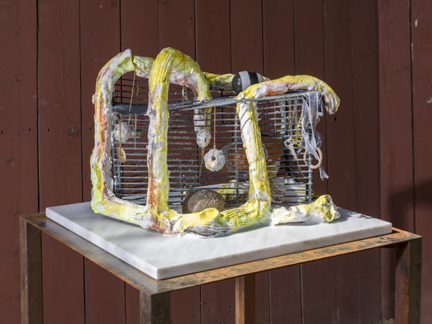 A metal cage is framed by a yellow and orange ceramic structure. Various found and crafted objects hang in and around the sculpture.