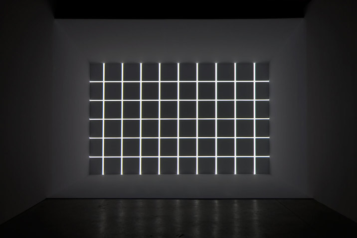 At the far wall of a grey room, there is a grid of lights on a dark grey background. 