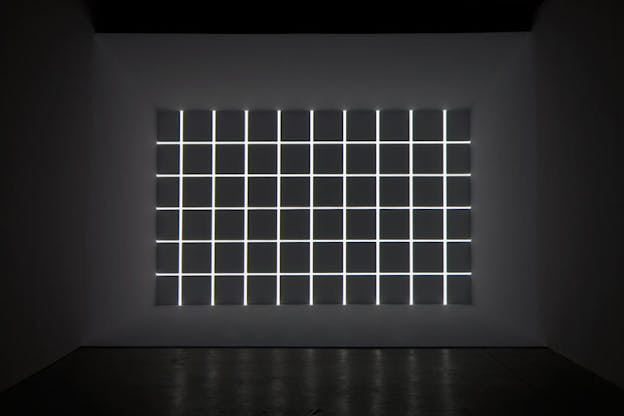 At the far wall of a grey room, there is a grid of lights on a dark grey background. 