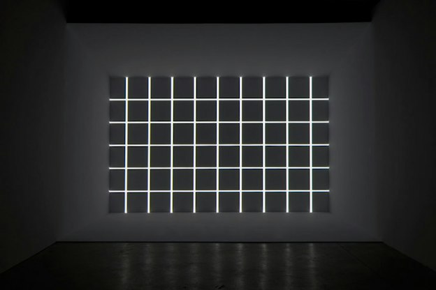 At the far wall of a grey room, there is a grid of lights on a dark grey background. 