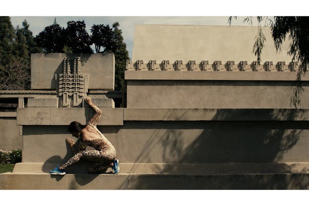 A figure in a cheetah print bodysuit with their back to the viewer kneels on an elevated marble platform of a beige wall while holding on to its top.