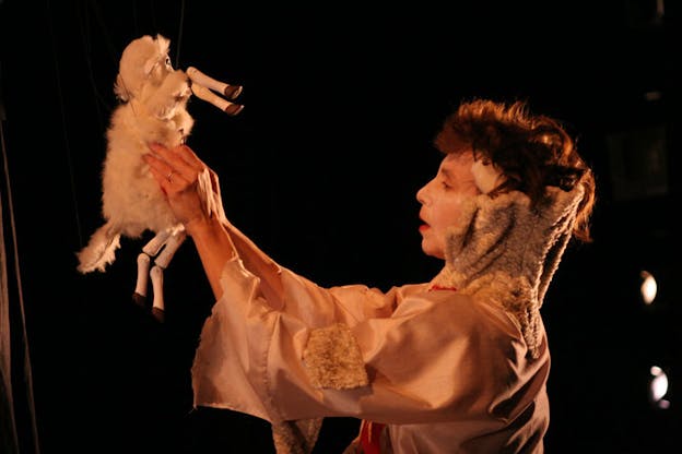 Against a black background, one performer facing left holds a sheep puppet hung from above. The performer wears a nude pink colored flare sleeve top with patches of teddy fabric. 