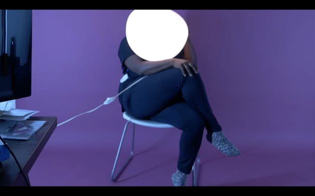 A performer sits with their legs crossed on a white plastic chair. They wear black leggings and grey spotted socks and their head is obscured by a white light orb. The wall behind them is purple. 