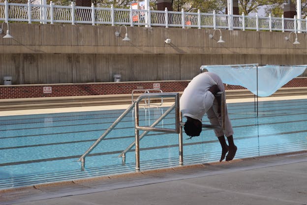 Autumn Knight slumps over a railing connected to the stairs leading into a pool. She wears a white t-shirt and kahki pants. Her feet are lifted and her head is turned down. To the left of her there is a structure in the pool made of metal cube frame with cloth attached to it, forming a bulbous shape. Beyond and above the pool there are trees and red brick columns. 