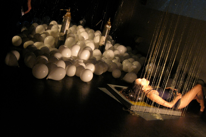 A tilted photograph of a figure on the far right lying down on a wooden replica of a beehive frame while being poured honey. Behind them on the left sit multiple white balloons.