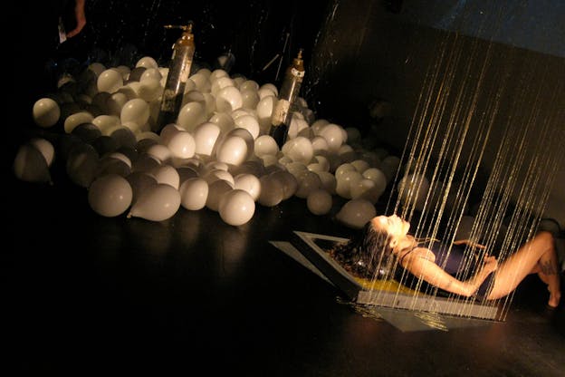 A tilted photograph of a figure on the far right lying down on a wooden replica of a beehive frame while being poured honey. Behind them on the left sit multiple white balloons.
