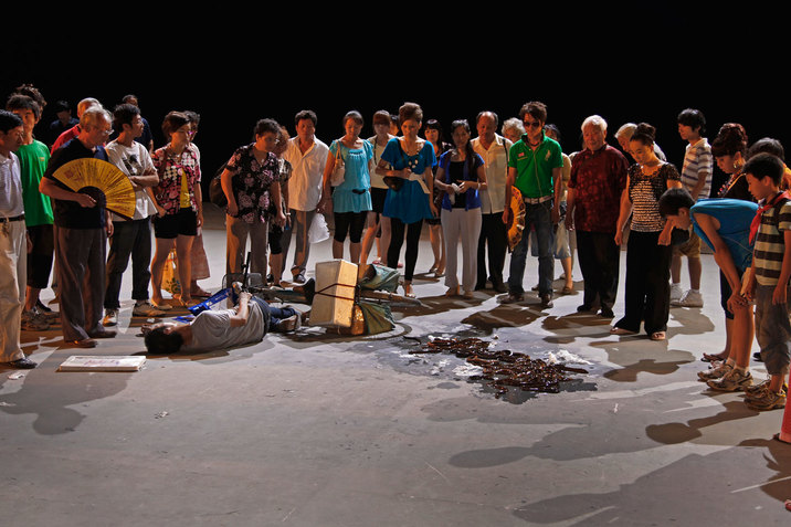 A performance still of numerous performers in various street clothes standing in a semi-circle around a person who appears to have fallen off their bike. This performer lays on their side and by their head, there is a white framed picture. To the side of them, many bronze colored, shiny wet snake like objects are scattered on the floor.  