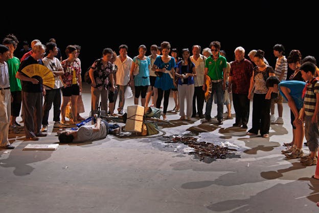 A performance still of numerous performers in various street clothes standing in a semi-circle around a person who appears to have fallen off their bike. This performer lays on their side and by their head, there is a white framed picture. To the side of them, many bronze colored, shiny wet snake like objects are scattered on the floor.  