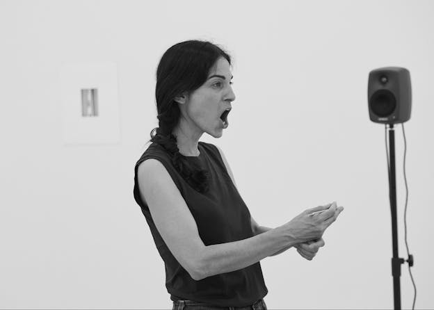 A black and white photograph of vocalist Inbal Hever in a gallery room with a black speaker in the background to her right and a couple of photos on the wall behind her. She is turned toward the right and her mouth is open as she vocalizes, her hands are raised in front of her body. She is wearing a black sleeveless shirt.