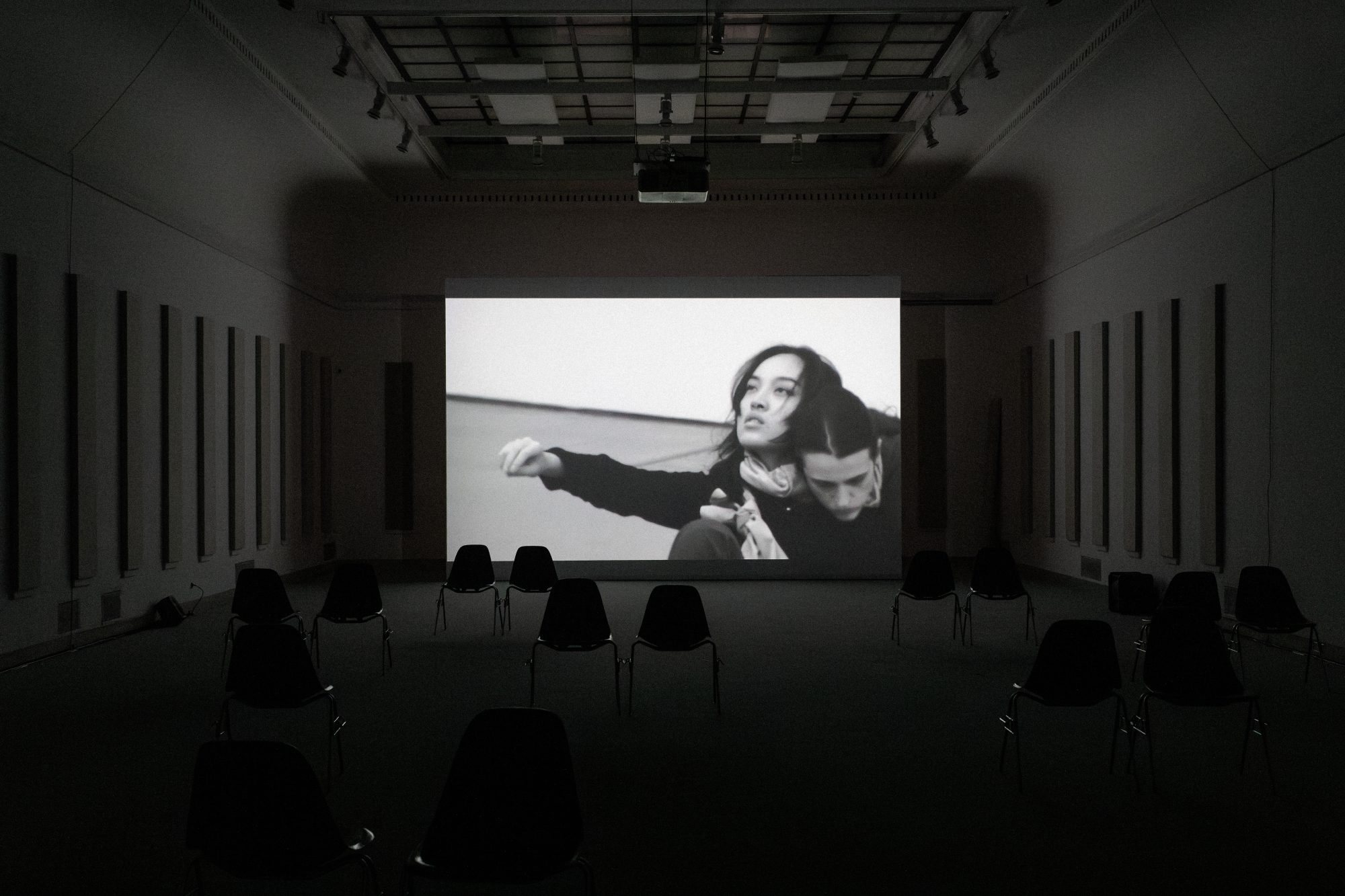 Media installation by Pan Daijing. In a dark exhibition space a black and white film is seeing playing on a large screen. In the video two figures, the artists and a collaborator, are seen in the corner of the screen with their bodies appearing to be intertwined with one another’s. Pan’s arm is seen outstretched out to her right. Within the exhibition space, on the left and right walls, are seen a series of tall and narrow speakers.