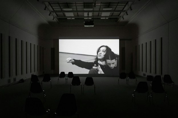 Media installation by Pan Daijing. In a dark exhibition space a black and white film is seeing playing on a large screen. In the video two figures, the artists and a collaborator, are seen in the corner of the screen with their bodies appearing to be intertwined with one another’s. Pan’s arm is seen outstretched out to her right. Within the exhibition space, on the left and right walls, are seen a series of tall and narrow speakers.