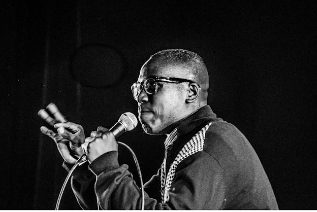 A black and white photograph of a performer gesturing with their right hand and holding a microphone up to their mouth with their left hand. They wear black glasses and a grey sweater with sections patterned with black and white repeating diamonds. The background is black. 