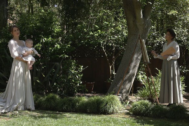 In a green garden, a person wearing a long skirt and blouse paints a person in a flowy white western-style dress holding a baby. 