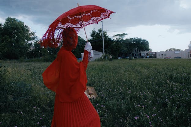 An artist stands in a field clad in a red pleated dress, lacy red facial covering, and white gloves, holding a red lace parasol over their head. They stand in profile with their head turned to the camera.