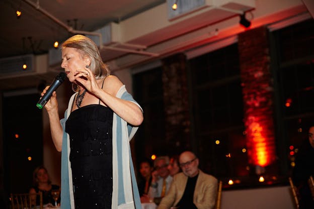 Photograph of a person with shoulder-length gray hair, a black sequined dress, a necklace and a blue scarf thrown on the arms singing in front of a microphone.