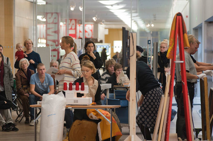 Backstrom speaks to an audience lined against a wall while in front of her, a person sits before a sewing machine holding an orange banner or sheet of fabric in their lap. 