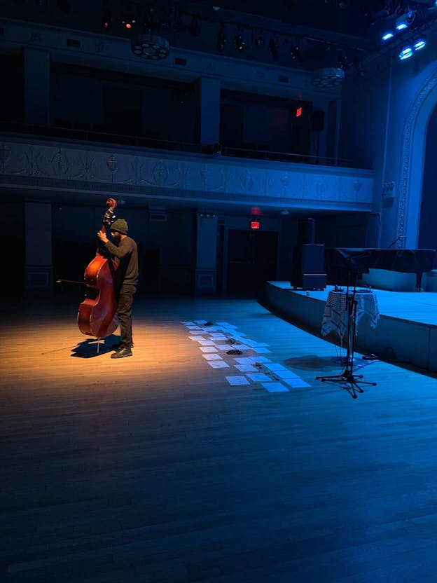 Luke Stewart plays his upright bass in a spotlight. The rest of the stage is lit by a soft blue light. Pieces of paper lie on the floor in three rows behind him.