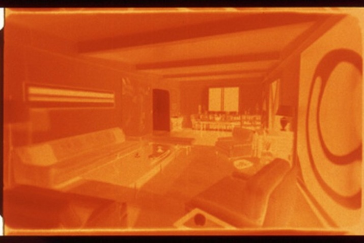 An orange hued image of a living room interior. On the left there is a couch and a big glass coffee table. On the right, a large painting of a circle hangs on the wall.