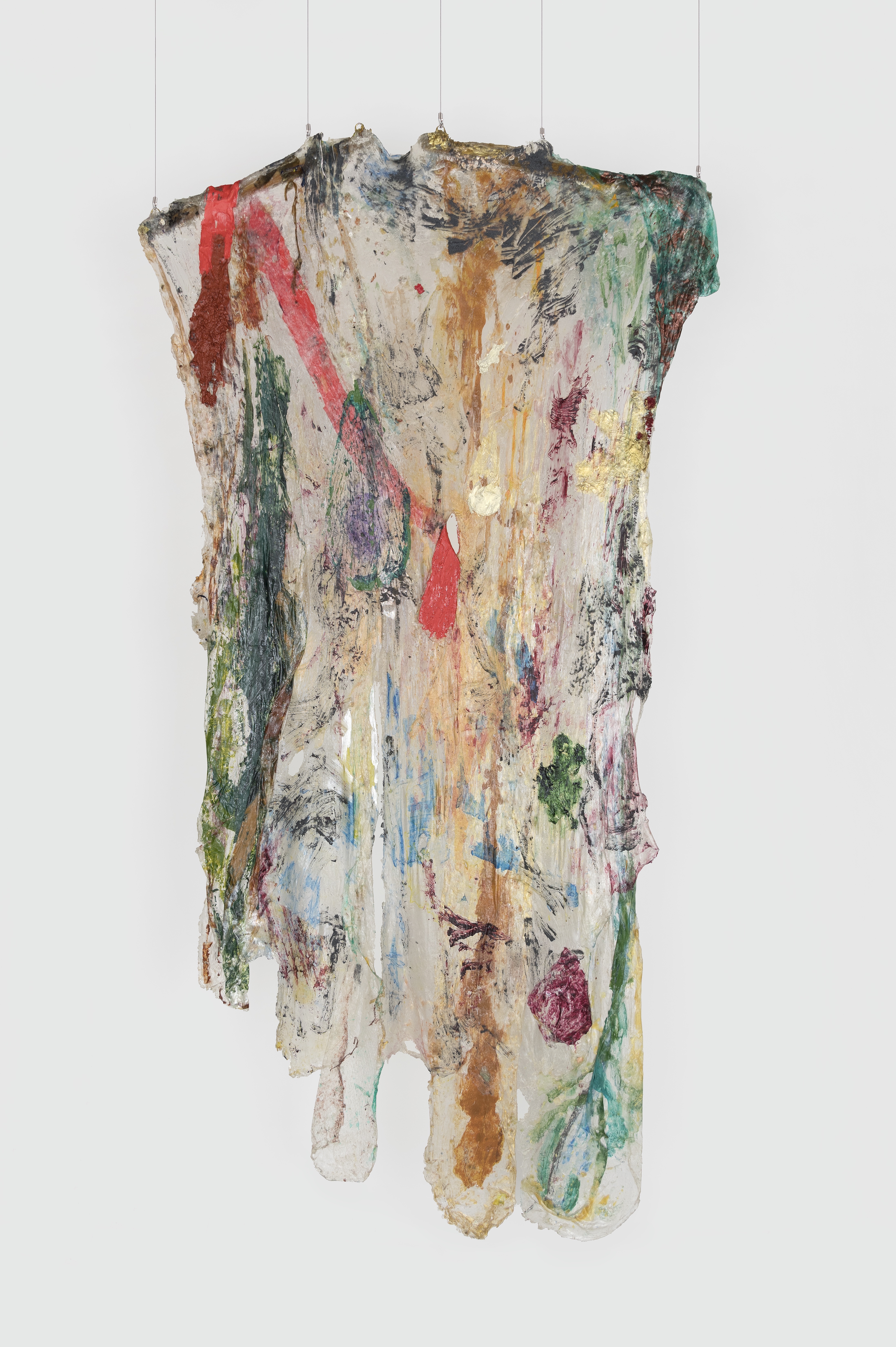 A hanging sculpture made of transparent plastic draped over two, short wooden rods, smeared with paint. The rods are tilted slightly, resembling the arms of clothes hanger. The plastic ends are cut in rounded forms. Dark green, red, brown, and magenta paint is visible on the plastic. A strip of red plastic at the top left hangs along the transparent piece, the bottom end coming through a hole in the center of the piece. 