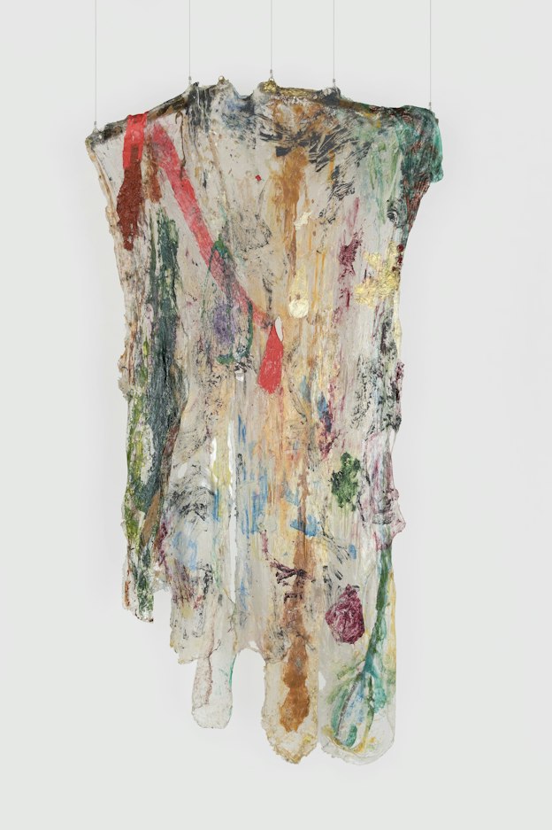 A hanging sculpture made of transparent plastic draped over two, short wooden rods, smeared with paint. The rods are tilted slightly, resembling the arms of clothes hanger. The plastic ends are cut in rounded forms. Dark green, red, brown, and magenta paint is visible on the plastic. A strip of red plastic at the top left hangs along the transparent piece, the bottom end coming through a hole in the center of the piece. 