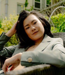  A low-angle portrait of a seated Haruna Lee, wearing a turquoise jacket shirt. Their head is supported by their right arm while their left arm lies relaxed.