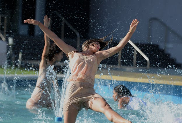 Three performers dance in a swimming pool outdoors in the sun, centered in a triangle with one dancer in the front in sharp focus, leaping out of the water, and two in the back, in softer focus. Paola Adorno sprawls with her body facing the camera with her torso tilted backward, head tossed to the right, and her arms spread in a Y. Her left leg is kicked forward, and she is wearing a light tan camisole with lace trim and loose tan lounge shorts. Carla Mendoza and Siul Valentín Medina dance in the background behind Adorno as sprinklers spout water in the foreground and middleground.