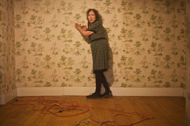 Performer looks uneasily at something in the distance as they lean against a wall decorated with sparse green tree wallpaper, in a dim yellow-lit unfurnished room. Rope and red extension cord are looped and tangled on the wooden floor. 