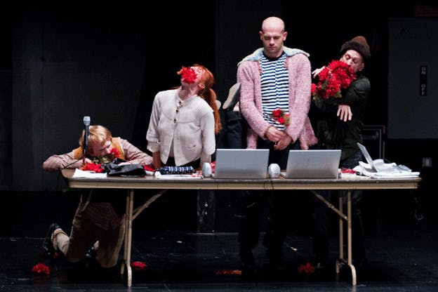 Four performers at a desk with three open laptops and other piled material, two performers' mouths are stuffed with red flowers from a bouquet a third performer is holding, while a fourth holds a flower in their hand and looks down at the laptops. 