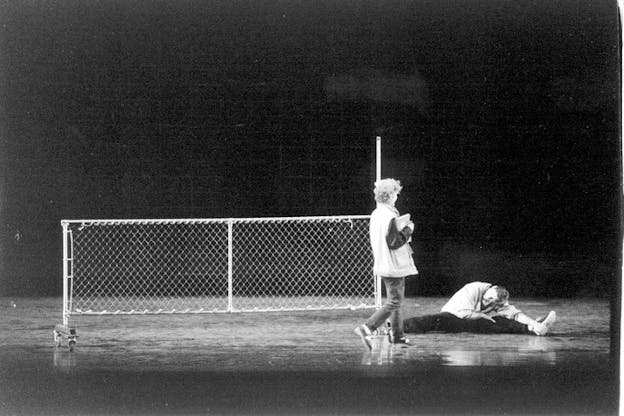A black and white performance still of two performers on a bare stage filled only with a small chain link fence. One performer sits on the floor and stretches over their left leg. The other performer walks up to them. Both performers wear casual street clothes. 