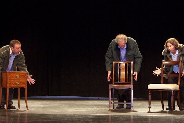 Photograph of three people similarly dressed, with the two on the right standing further from the one on the left. All three of them stand in front of furniture, the two on the right with chairs and the figure on the left with a nightstand. All three figures have their palms open and extended towards the furniture. 