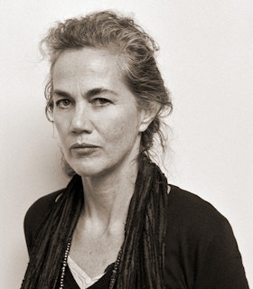A black and white portrait of Joan Waltemath angled against a wide wall. While her body is angeled to the side, she faces the camera directly. Her hair is pulled into a low bun and she wears a black shirt and thin black textured scarf. 