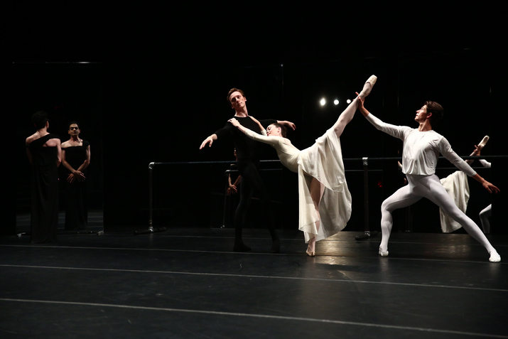A ballet dancer dressed in black supports a ballet dancer dressed in a white long skirt who lifts their leg upwards and holds unto to the shoulders of the other. A third ballet dancer, dressed in white behind the middle one, kneels and turns their front leg while holding the middle dancers upward leg.