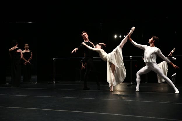 A ballet dancer dressed in black supports a ballet dancer dressed in a white long skirt who lifts their leg upwards and holds unto to the shoulders of the other. A third ballet dancer, dressed in white behind the middle one, kneels and turns their front leg while holding the middle dancers upward leg.