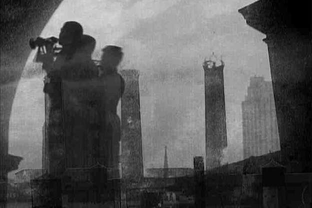 A black and white, slightly blurry video still of three people huddled together on the left side facing out towards the left, beyond the image. In the background, the tops of buildings are visible.