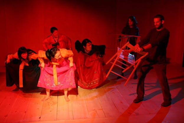 Four people dressed in traditional Koreans wear are in a squat position while making angry expressions to the two people dressed in brown-toned clothes on their right. The two people each holds a wooden folding chair in front of them. The set has wood floors and walls lit by fluorescent red lights.