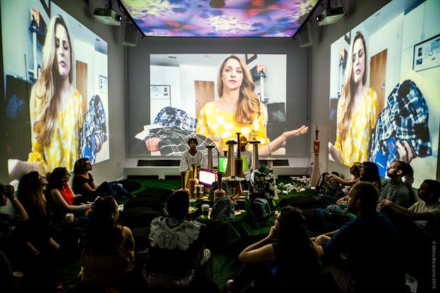 People sit in a dark room with images are projected onto three walls and the ceiling. Tei Blow and Sean McElroy kneel with their eyes closed at the far end of the room, facing the camera with a collection of tall, long necked vessels in front of them. Projected on the three visible walls is an image of a woman with long hear wearing a yellow patterned blouse, holding two flannel shirts, and speaking to the camera. Colorful geometric patterns are being projected onto the ceiling. Blow and McElroy are both wearing loose white tunics and golden wreaths on their heads.
