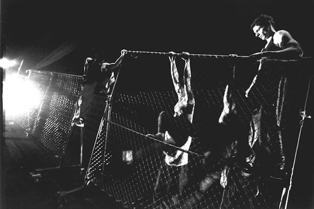 A black and white performance still of several performers climbing over a chain link fence in an otherwise bare space. A bright light shines from the left corner of the space. 