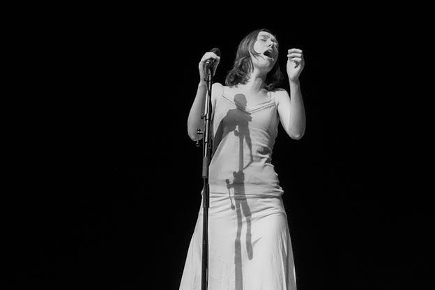 Black and white photograph of a person dressed in a long dress with neck-length straight hair singing in front of a microphone with closed eyes.
