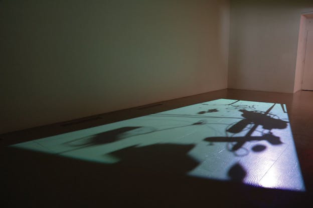 In a bare, grey room, a blue light is projected onto the floor. The light is interrupted by numerous, differently angled and sized shadows of a telephone pole, various wires, a small bicycle, and a street light. 