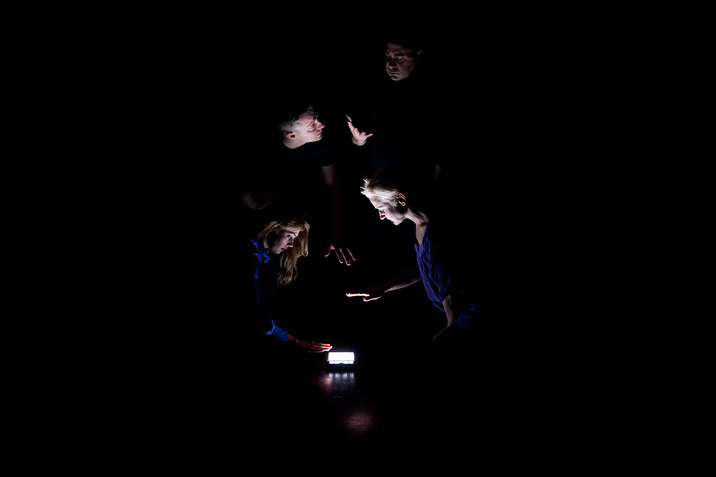 On a black-lit stage four persons huddle around an LED light placed on the floor, two kneeling and holding their palms over the light and two standing and gesturing to each other. 
