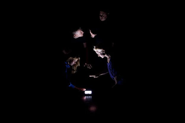 On a black-lit stage four persons huddle around an LED light placed on the floor, two kneeling and holding their palms over the light and two standing and gesturing to each other. 