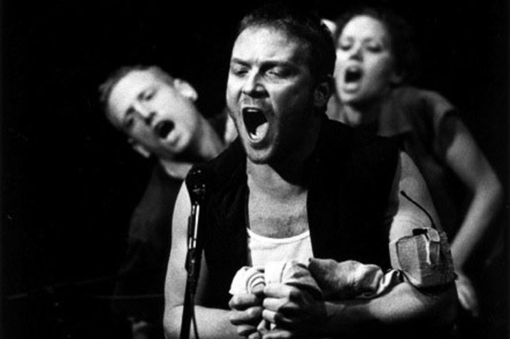 A black and white photograph of three performers with anguished, open-mouthed, expressions standing in front of a black background. One person in the foreground appears to speak into a microphone as they tightly clench fabric in their hands. They are wearing a white shirt and a dark vest. Behind them, two other performers lean forwards.