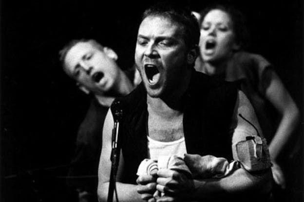 A black and white photograph of three performers with anguished, open-mouthed, expressions standing in front of a black background. One person in the foreground appears to speak into a microphone as they tightly clench fabric in their hands. They are wearing a white shirt and a dark vest. Behind them, two other performers lean forwards.